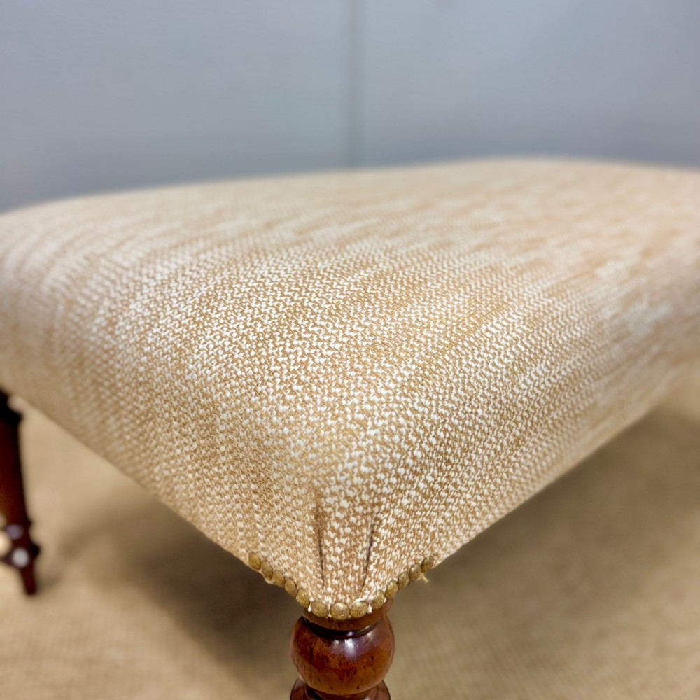 An oblong footstool upholstered in a neutral textured cream fabric supported by four turned warm brown timber legs. View III