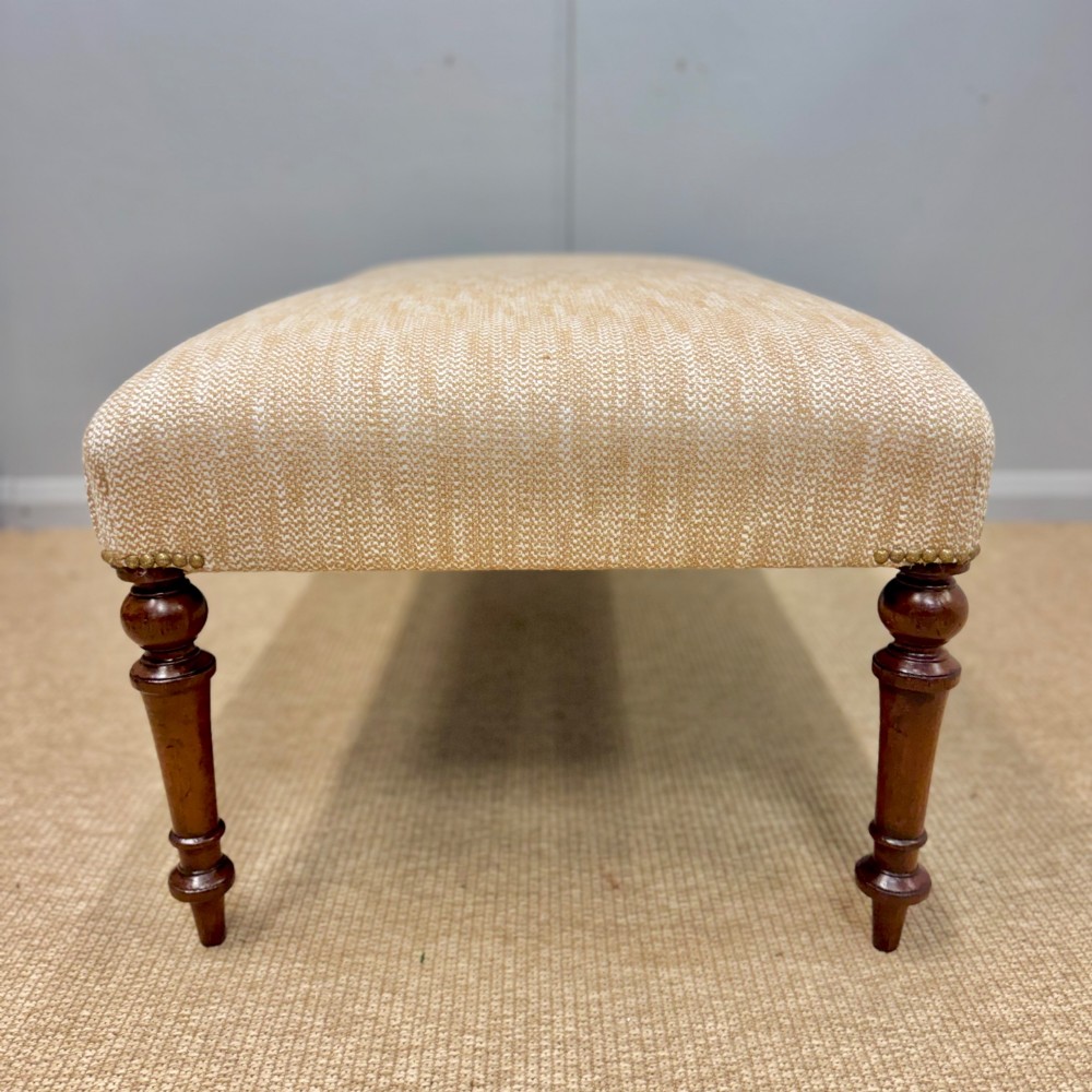 An oblong footstool upholstered in a neutral textured cream fabric supported by four turned warm brown timber legs. View IV