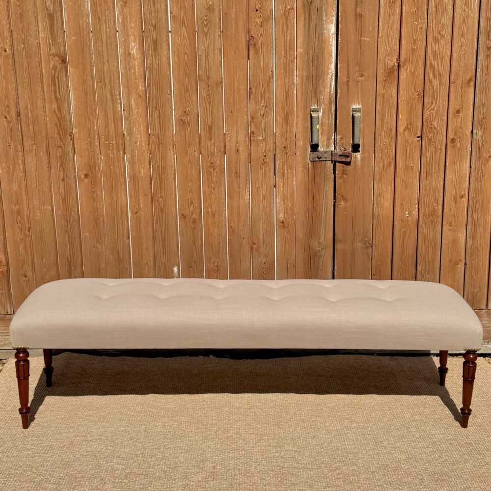 A long 185cm upholstered stool featuring a beige button-tufted linen top set on four turned dark wood legs.
