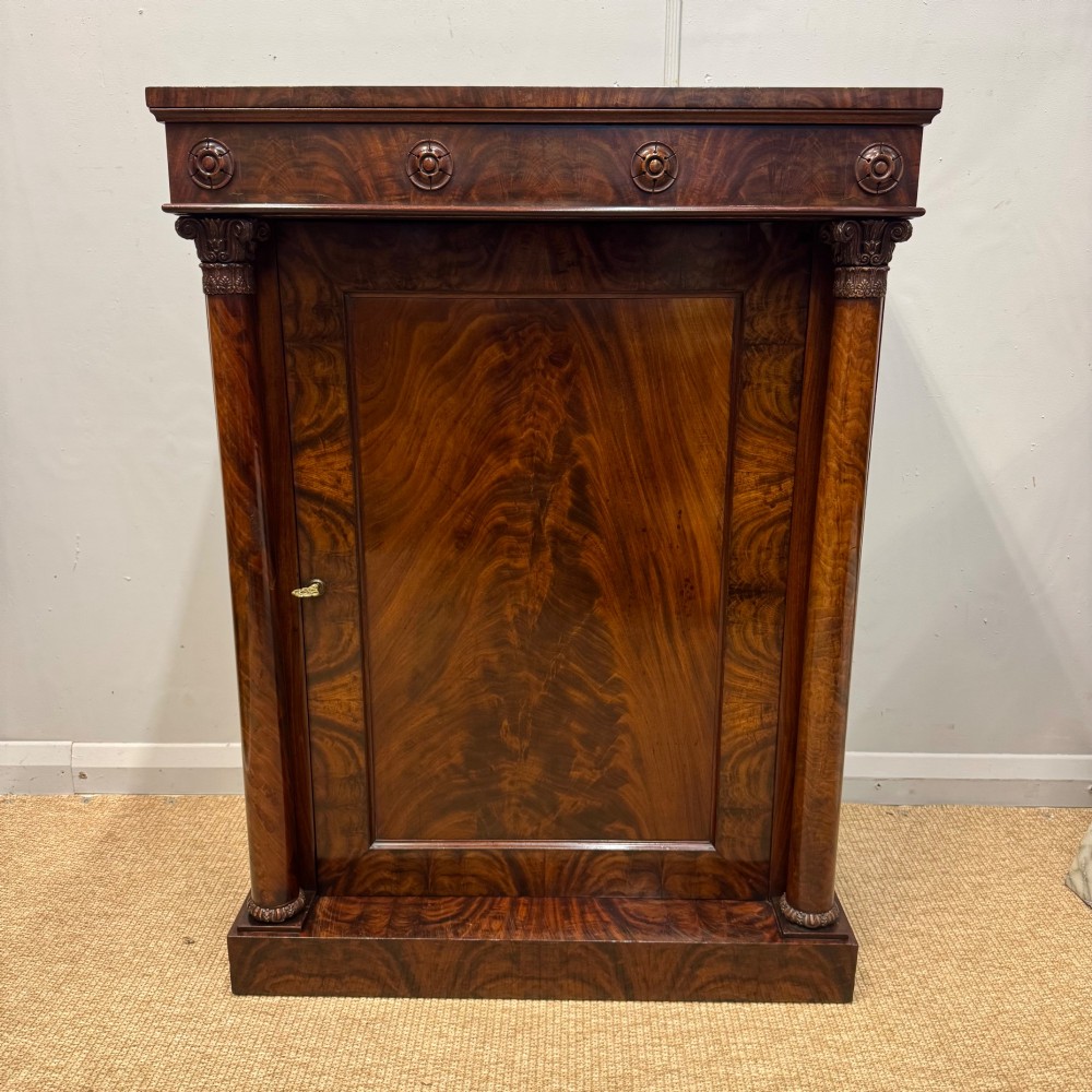 A Late Regency flame mahogany pier cabinet featuring carved rosette medallions on the upper frieze, flanking side columns with carved capitals, and a single solid wood door.