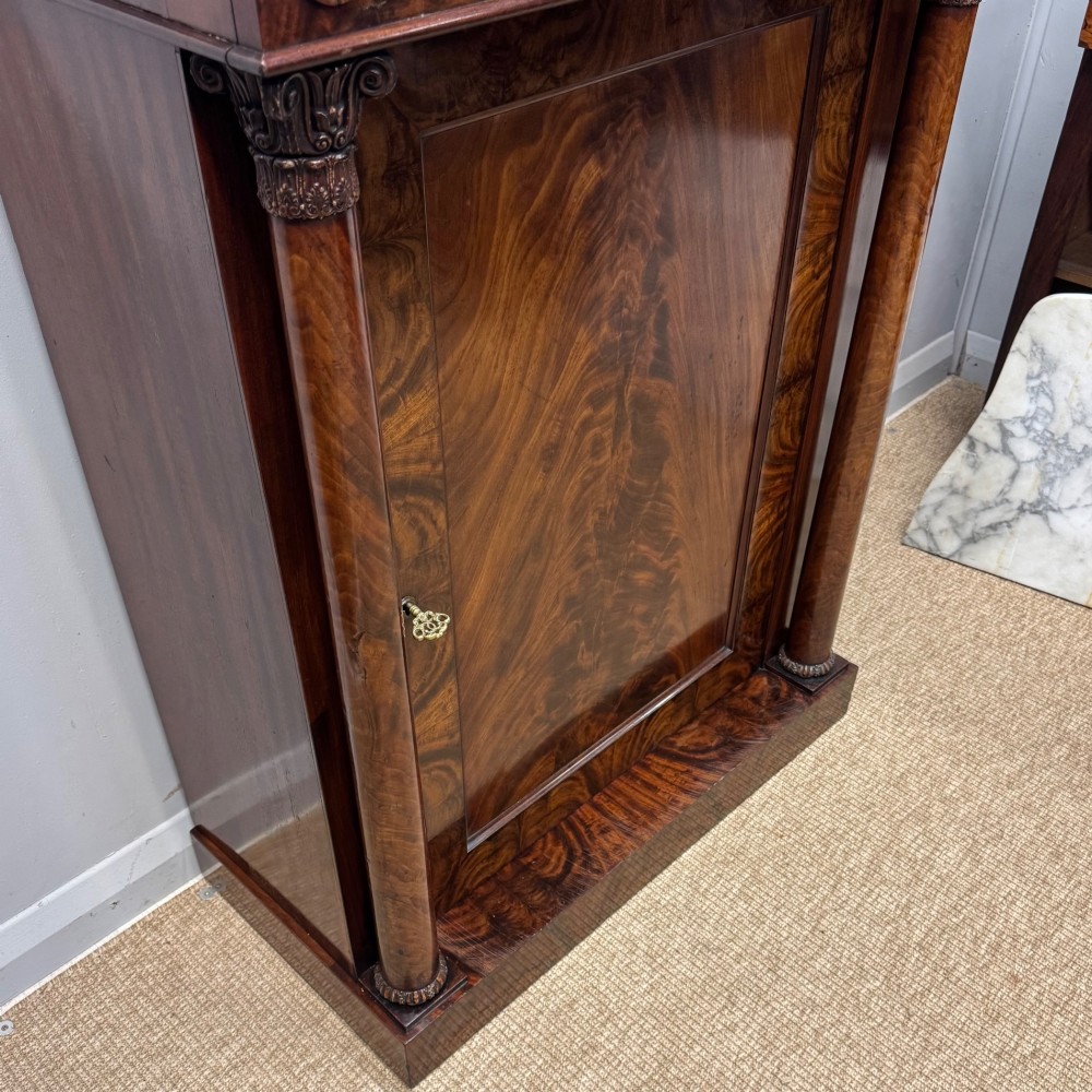A Late Regency flame mahogany pier cabinet featuring carved rosette medallions on the upper frieze, flanking side columns with carved capitals, and a single solid wood door. View III