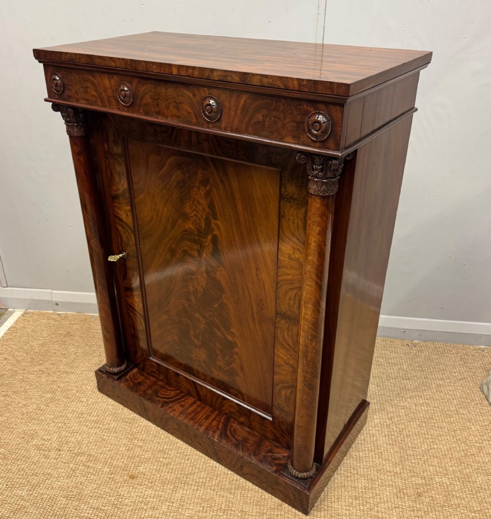 A Late Regency flame mahogany pier cabinet featuring carved rosette medallions on the upper frieze, flanking side columns with carved capitals, and a single solid wood door. View V