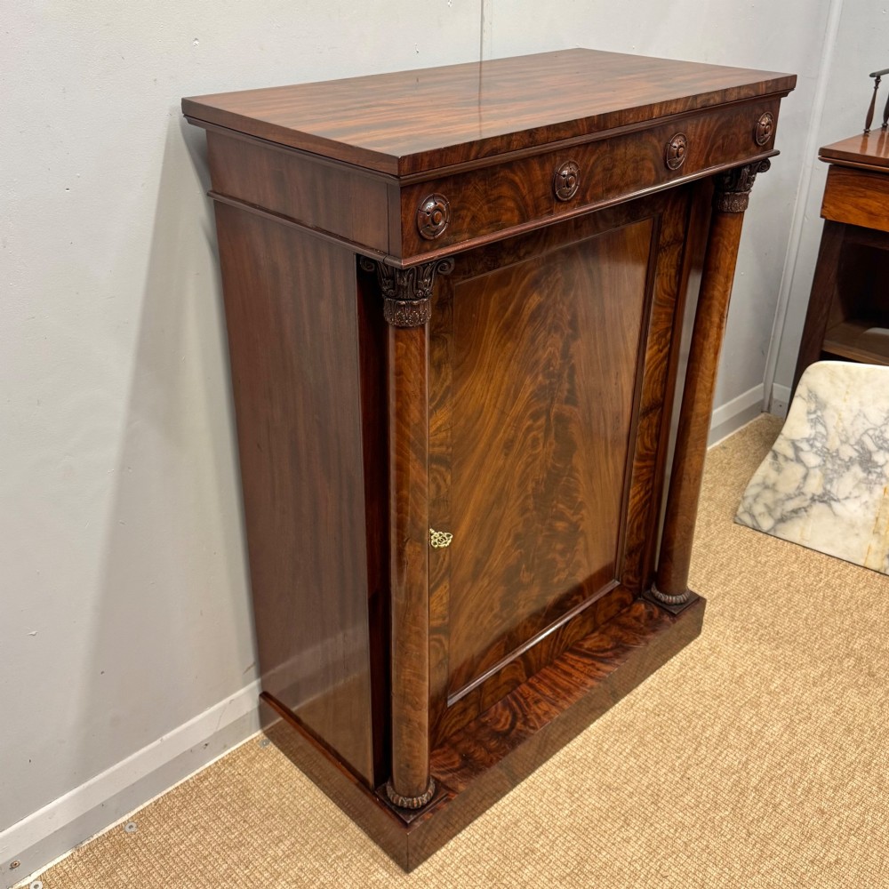 A Late Regency flame mahogany pier cabinet featuring carved rosette medallions on the upper frieze, flanking side columns with carved capitals, and a single solid wood door. View VI