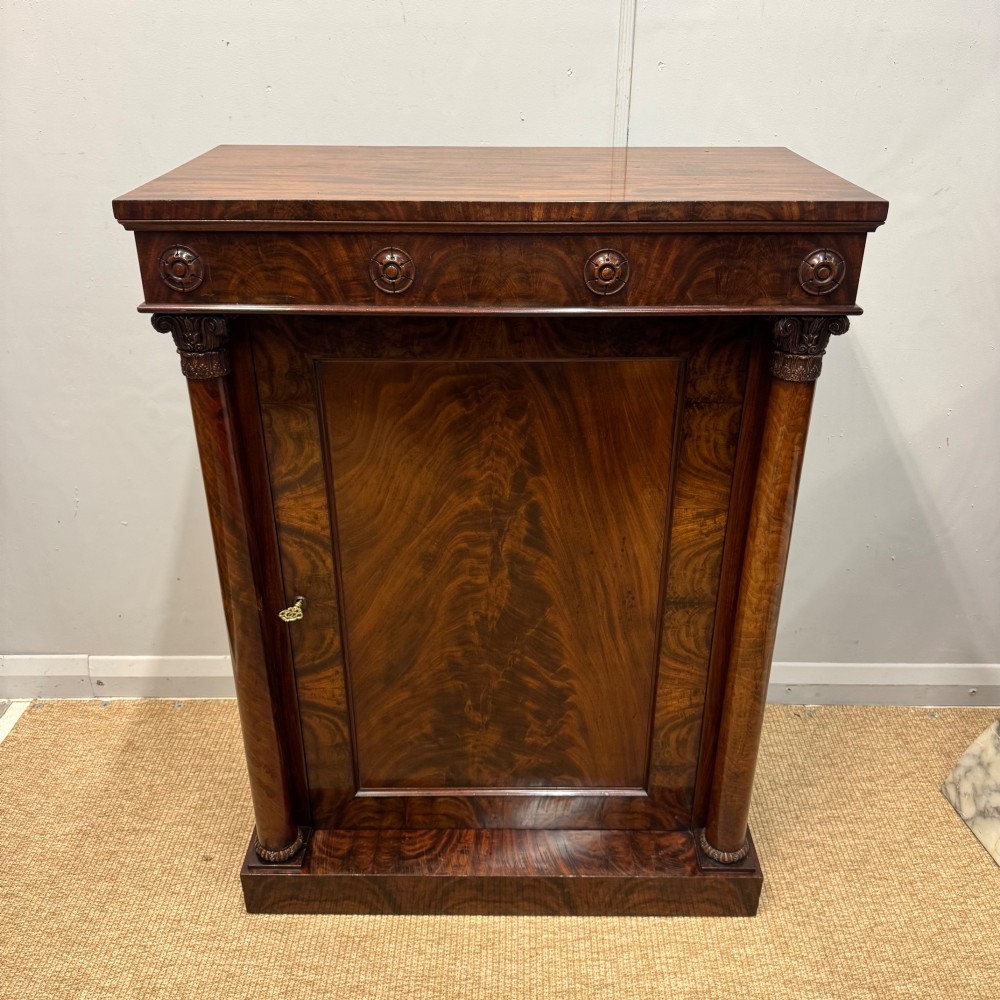A Late Regency flame mahogany pier cabinet featuring carved rosette medallions on the upper frieze, flanking side columns with carved capitals, and a single solid wood door. View VII