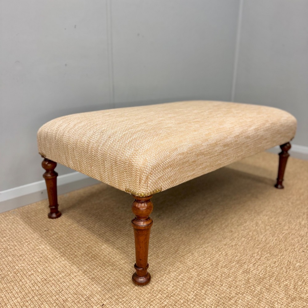 A rectangular upholstered footstool covered in beige chenille fabric, featuring brass stud trim at the corners and four turned wooden legs.