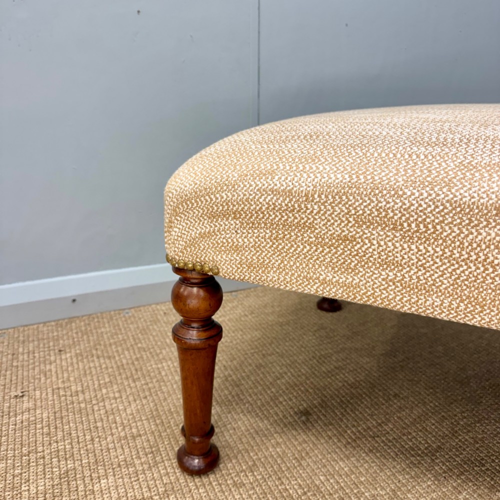 A rectangular upholstered footstool covered in beige chenille fabric, featuring brass stud trim at the corners and four turned wooden legs. View V