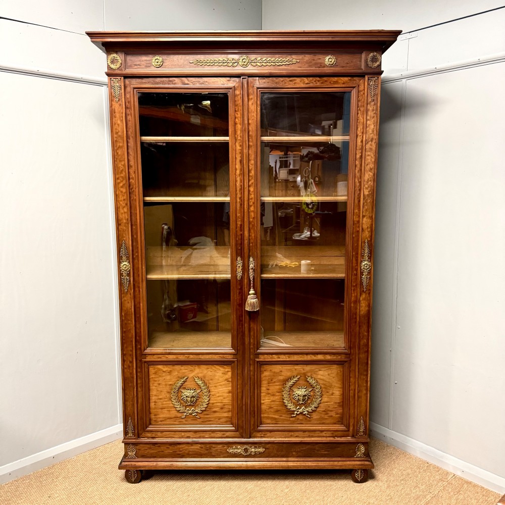 A tall French Empire style warm brown timber bookcase featuring two glass-fronted doors, gilt brass laurel wreath mounts, and adjustable interior shelves. View II