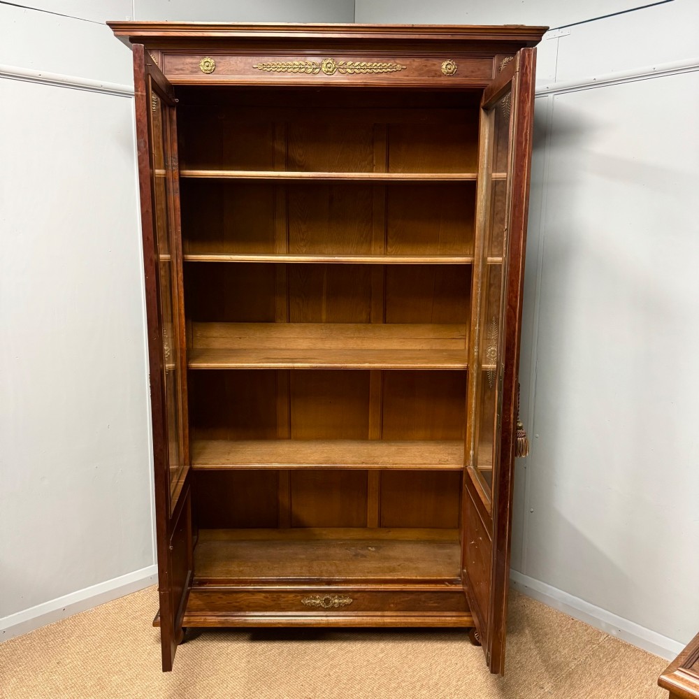A tall French Empire style warm brown timber bookcase featuring two glass-fronted doors, gilt brass laurel wreath mounts, and adjustable interior shelves. View IV