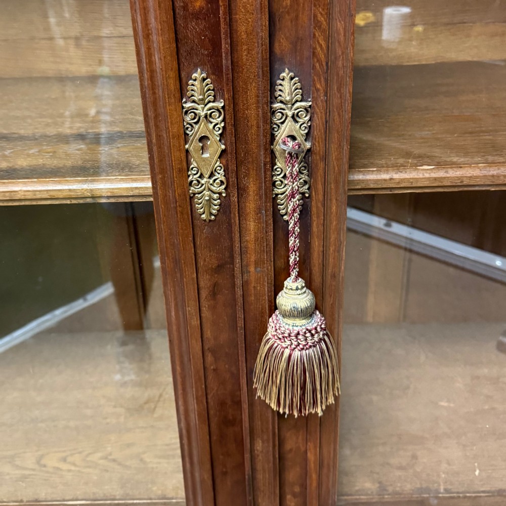 A tall French Empire style warm brown timber bookcase featuring two glass-fronted doors, gilt brass laurel wreath mounts, and adjustable interior shelves. View VI