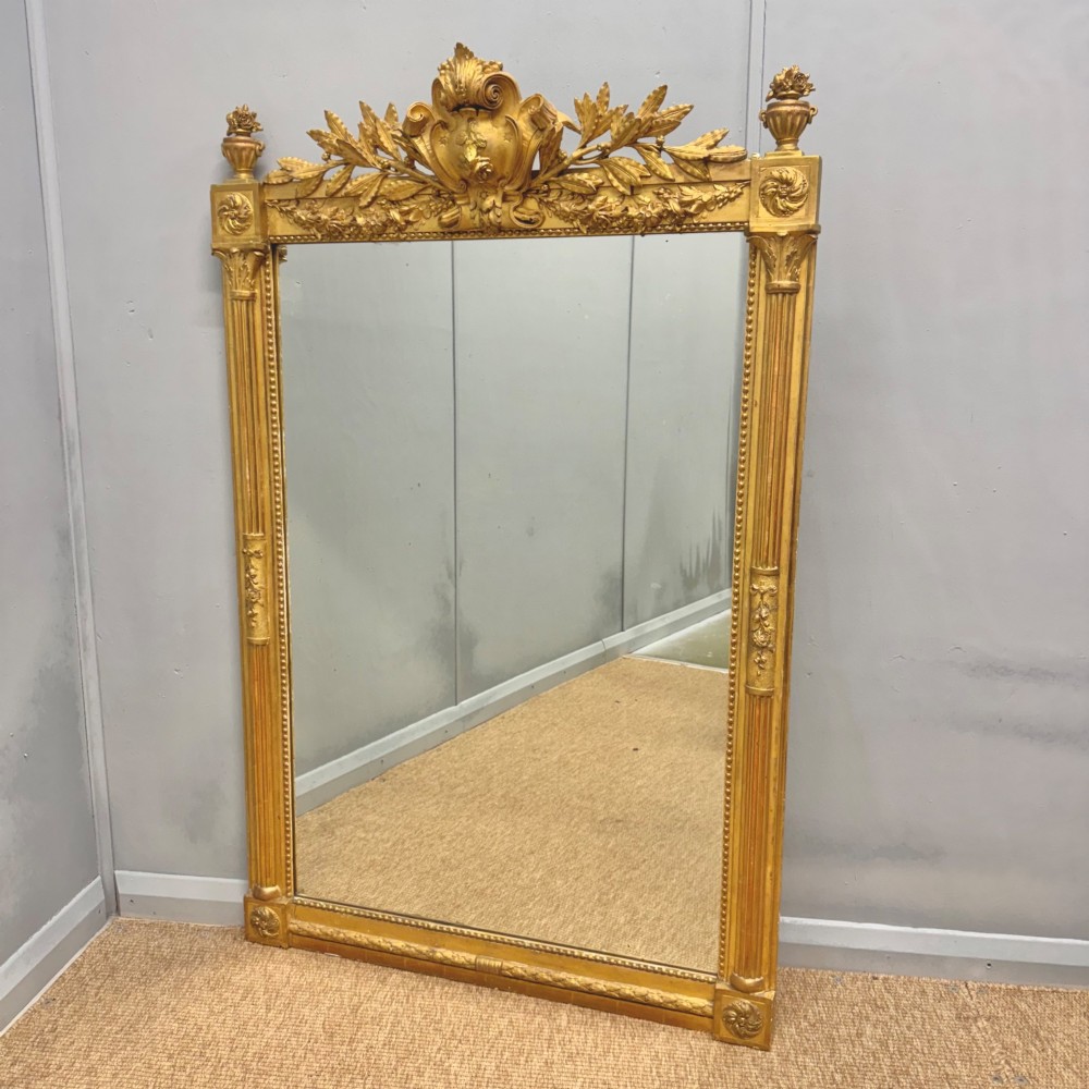 A large rectangular French Louis XVI style water gilded mirror featuring fluted side pilasters and an elaborate carved crest with laurel leaves and urn finials.