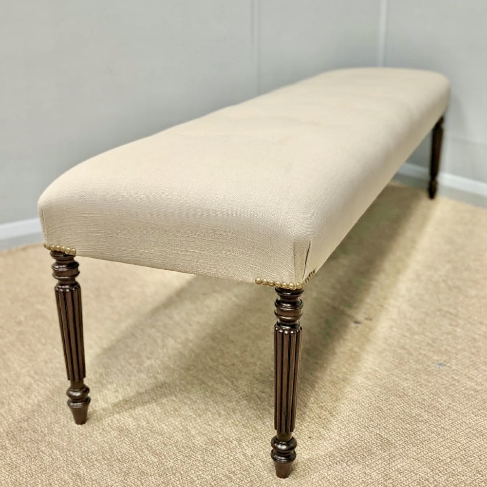 A long upholstered bench in neutral fabric featuring brass stud trim and dark fluted wooden legs.