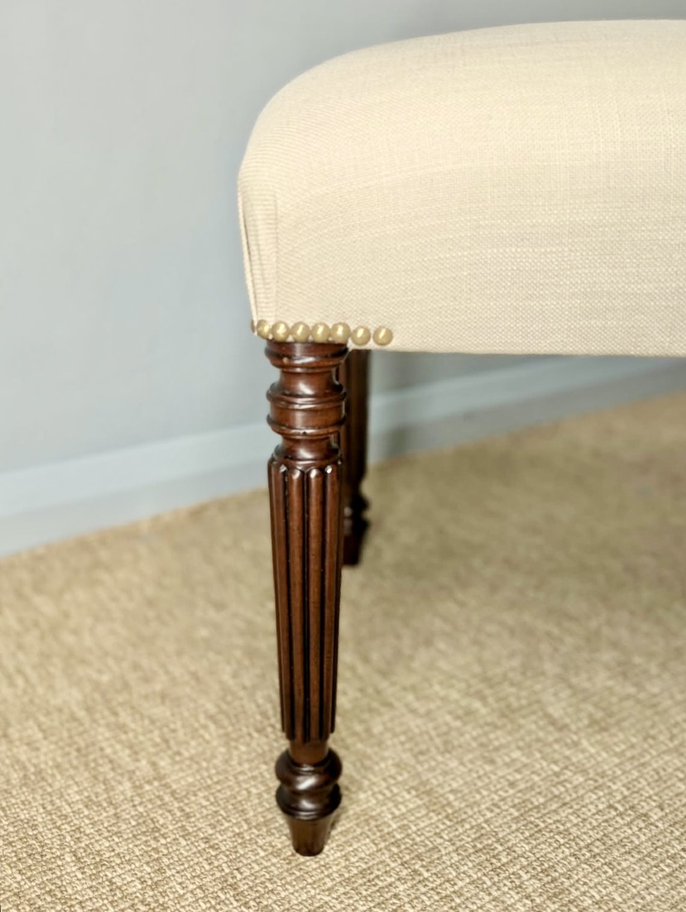 A long upholstered bench in neutral fabric featuring brass stud trim and dark fluted wooden legs. View II