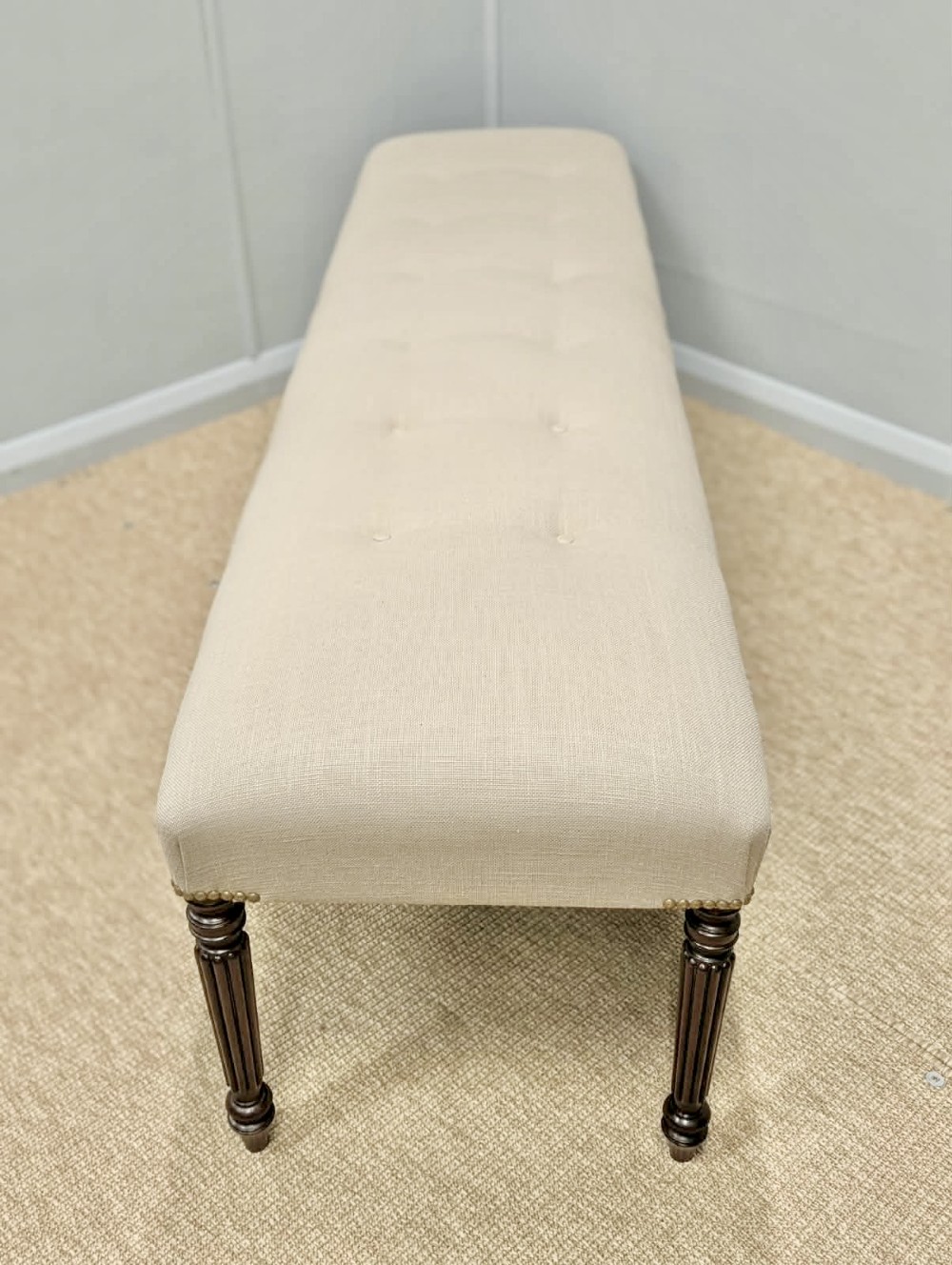 A long upholstered bench in neutral fabric featuring brass stud trim and dark fluted wooden legs. View IV