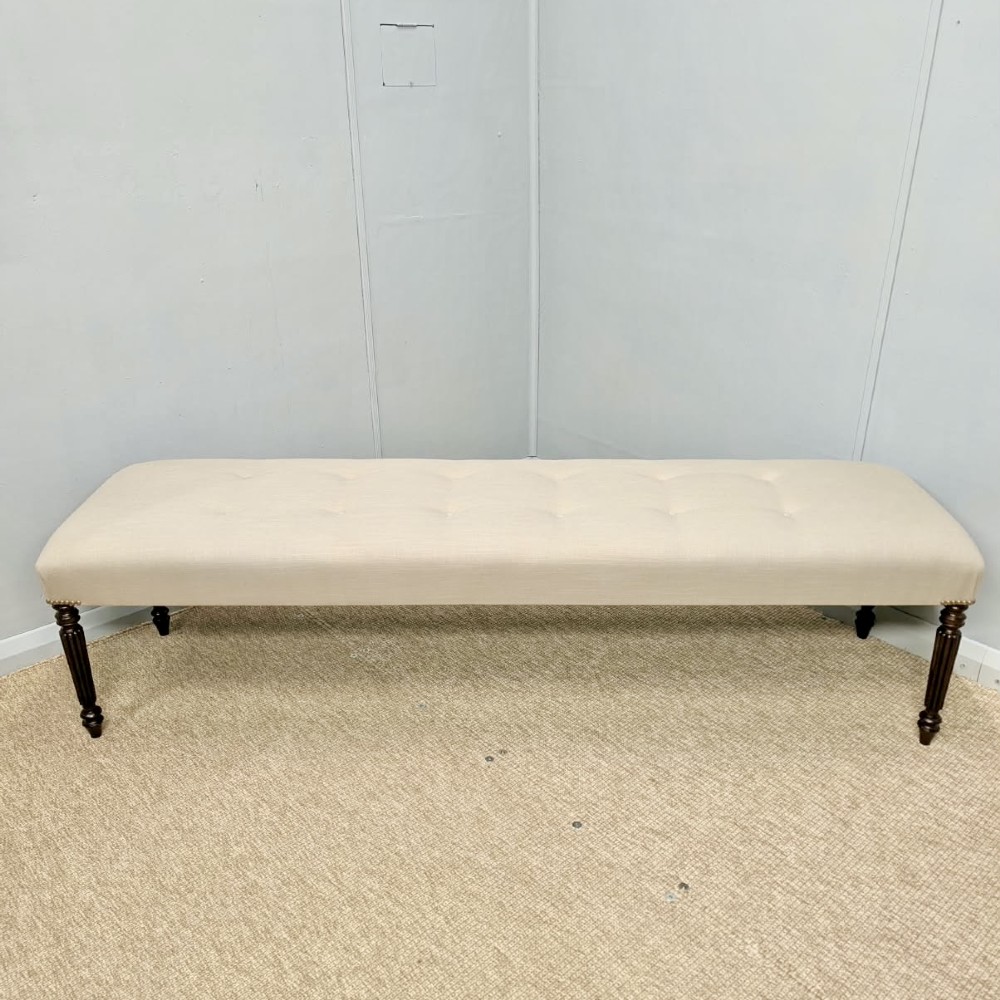 A long upholstered bench in neutral fabric featuring brass stud trim and dark fluted wooden legs. View V