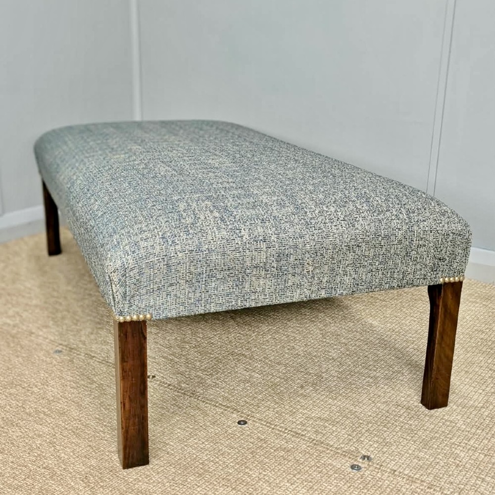 A large rectangular upholstered footstool covered in blue woven fabric with dark square wooden legs and brass stud detailing.