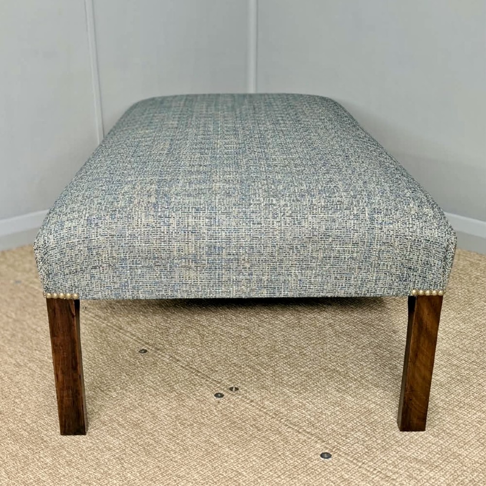 A large rectangular upholstered footstool covered in blue woven fabric with dark square wooden legs and brass stud detailing. View IV