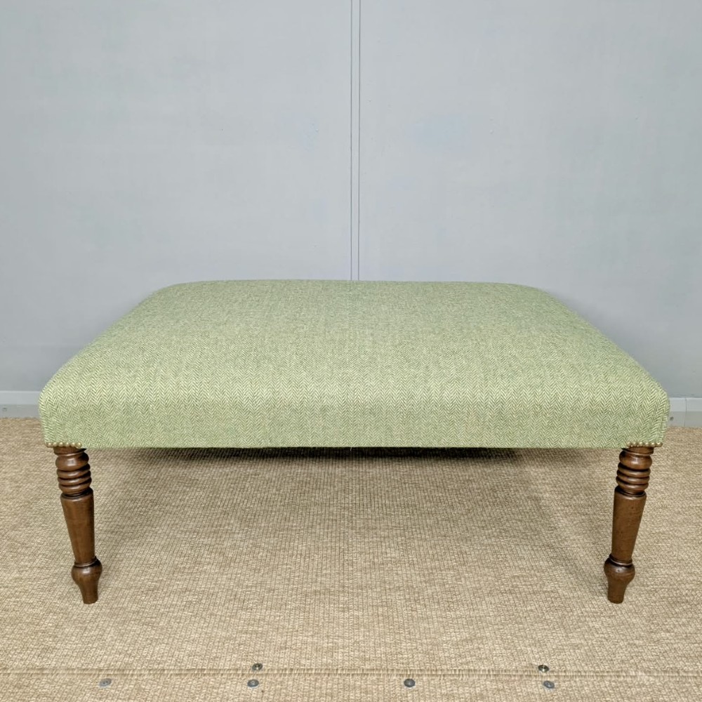 A square low footstool upholstered in sage green herringbone wool tweed with four turned dark wood legs and brass stud detailing. View II