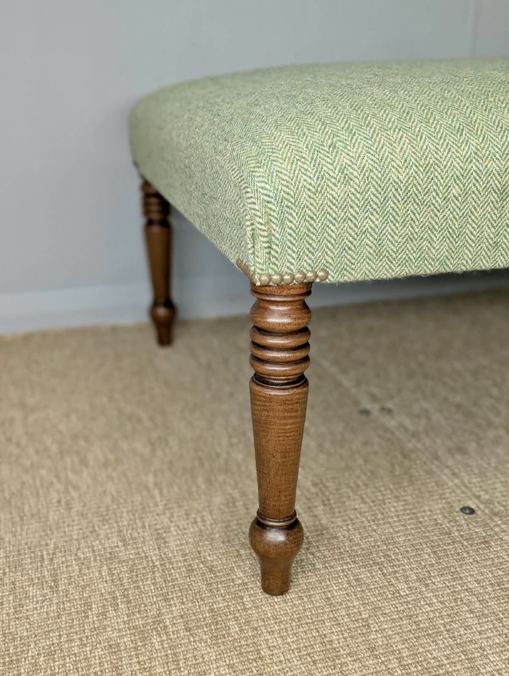 A square low footstool upholstered in sage green herringbone wool tweed with four turned dark wood legs and brass stud detailing. View III