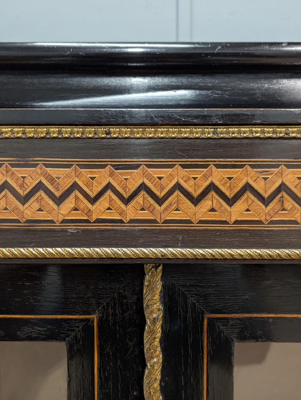 A black ebonised two-door glazed cabinet featuring a moulded caddy top, brass ormolu corner mounts, and a band of geometric marquetry inlay. View V