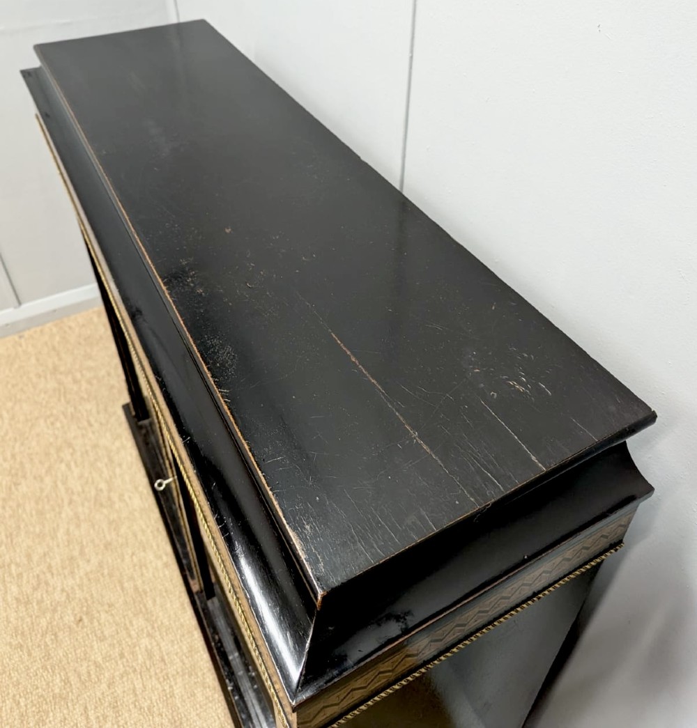 A black ebonised two-door glazed cabinet featuring a moulded caddy top, brass ormolu corner mounts, and a band of geometric marquetry inlay. View VIII
