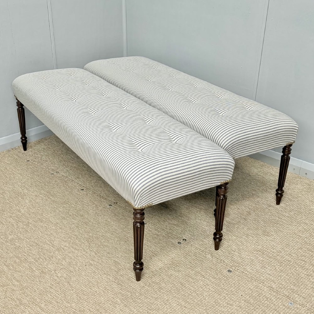 A pair of long upholstered benches featuring grey and white striped ticking fabric with deep button tufting set upon dark turned and fluted timber legs.