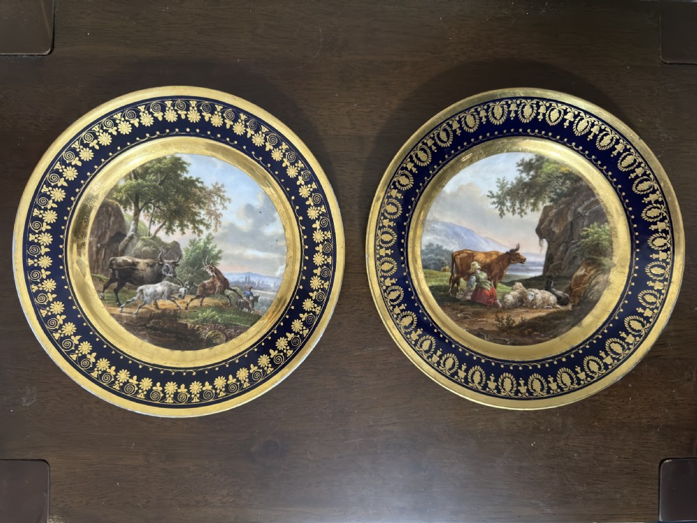 A pair of circular French Empire porcelain cabinet plates featuring hand-painted pastoral scenes surrounded by heavy gilded borders on cobalt blue.