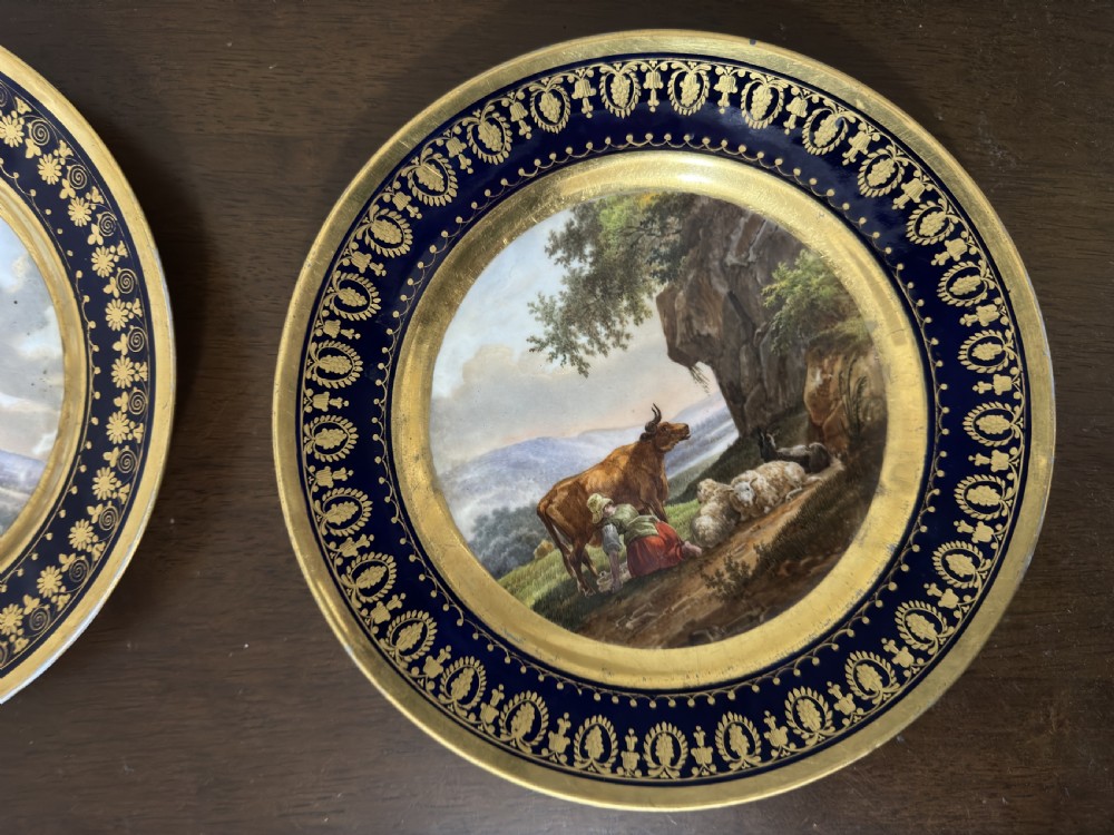 A pair of circular French Empire porcelain cabinet plates featuring hand-painted pastoral scenes surrounded by heavy gilded borders on cobalt blue. View V