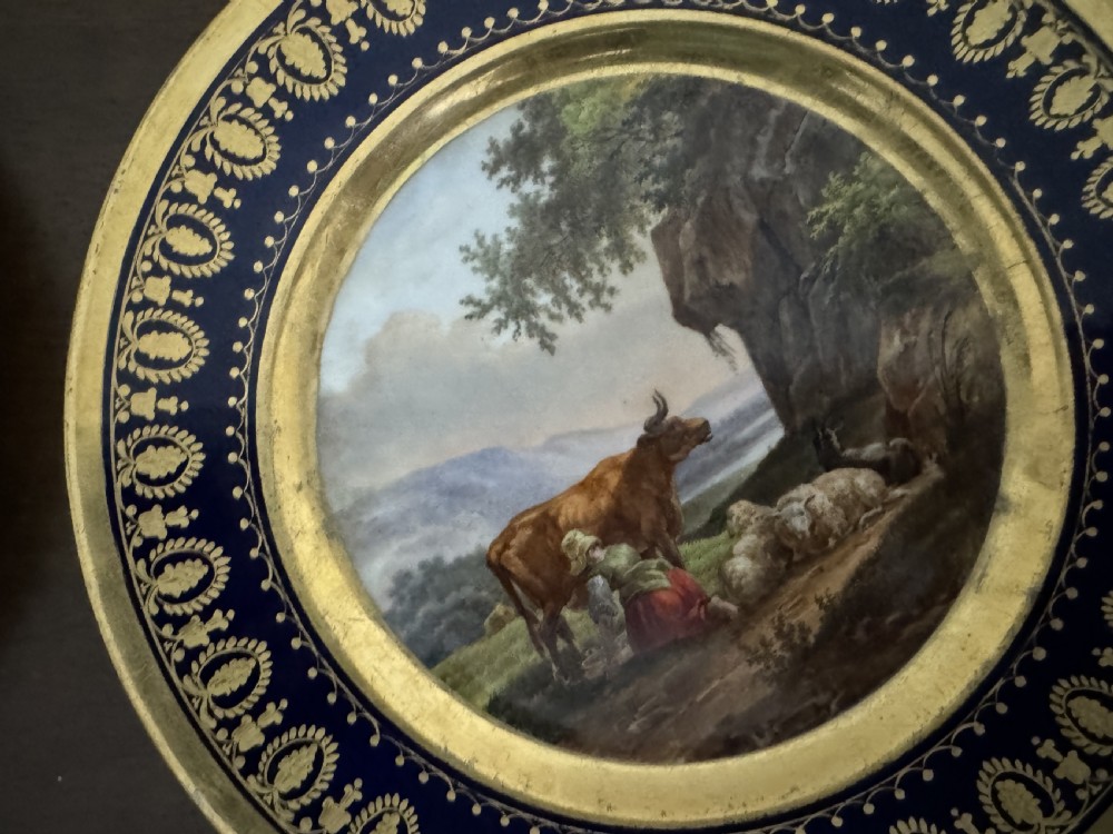 A pair of circular French Empire porcelain cabinet plates featuring hand-painted pastoral scenes surrounded by heavy gilded borders on cobalt blue. View VII