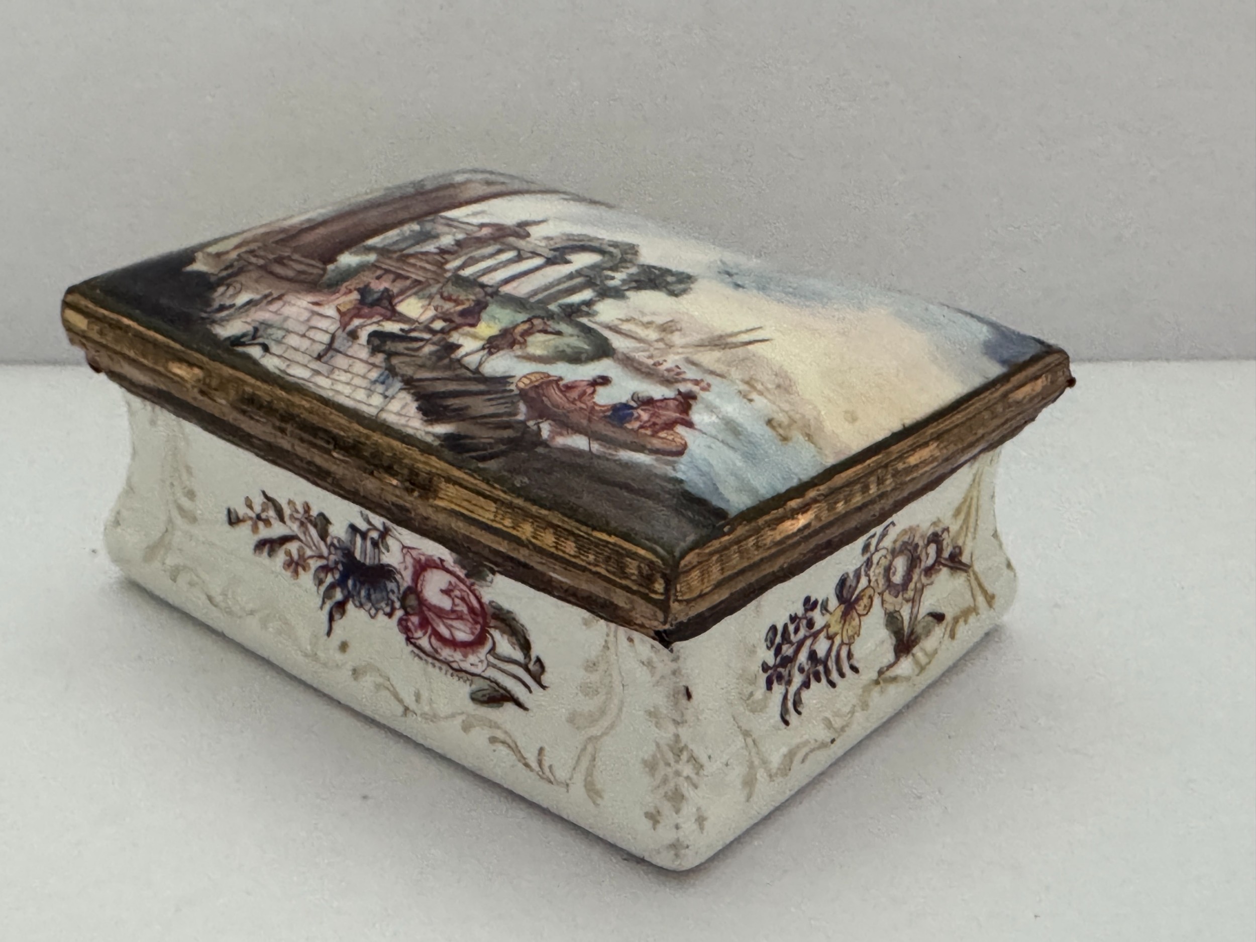 A rectangular 18th-century South Staffordshire enamel snuff box featuring a hand-painted lid with classical ruins, floral motifs on a white body, and gilded metal mounts.