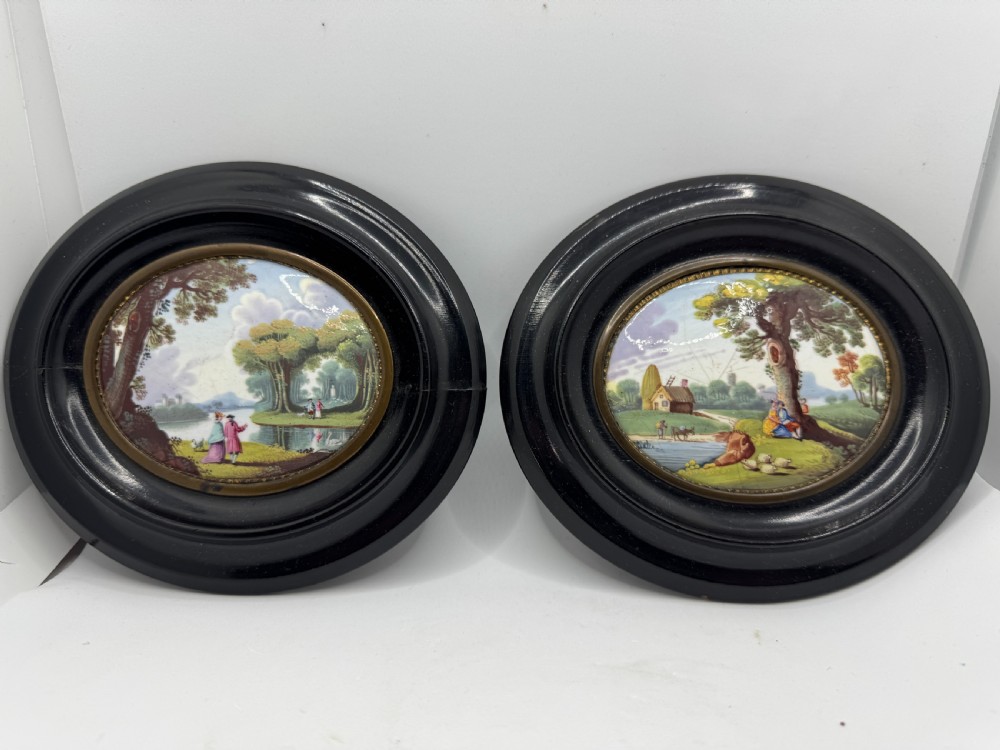 A pair of oval 18th-century enamel plaques featuring hand-painted pastoral landscape scenes set within black ebonised frames and brass bezels.