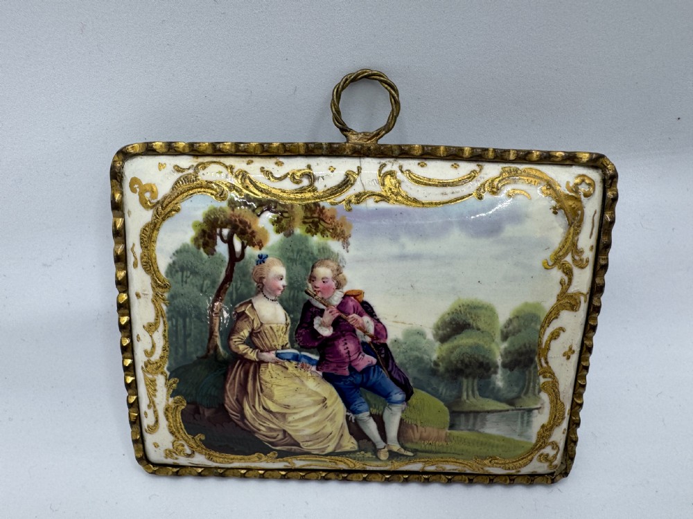 An eighteenth-century rectangular South Staffordshire enamel plaque depicting a hand-painted pastoral scene surrounded by gilt scrollwork and mounted in a metal frame with a hanging loop.