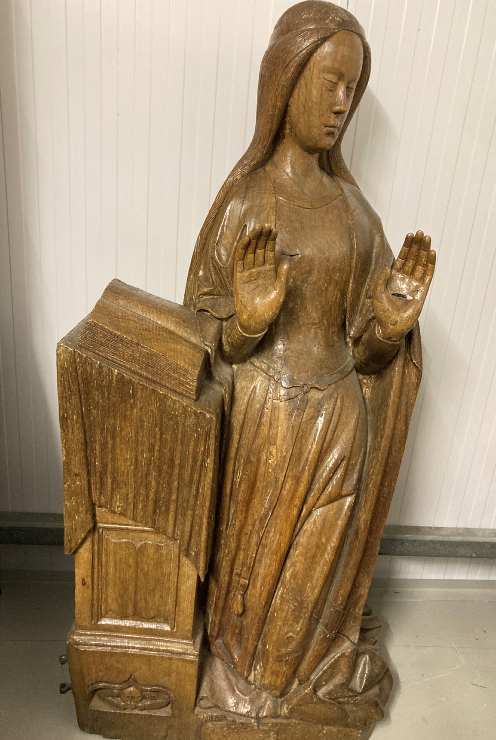A carved oak figure of the Virgin Mary standing beside a prayer desk with raised hands.