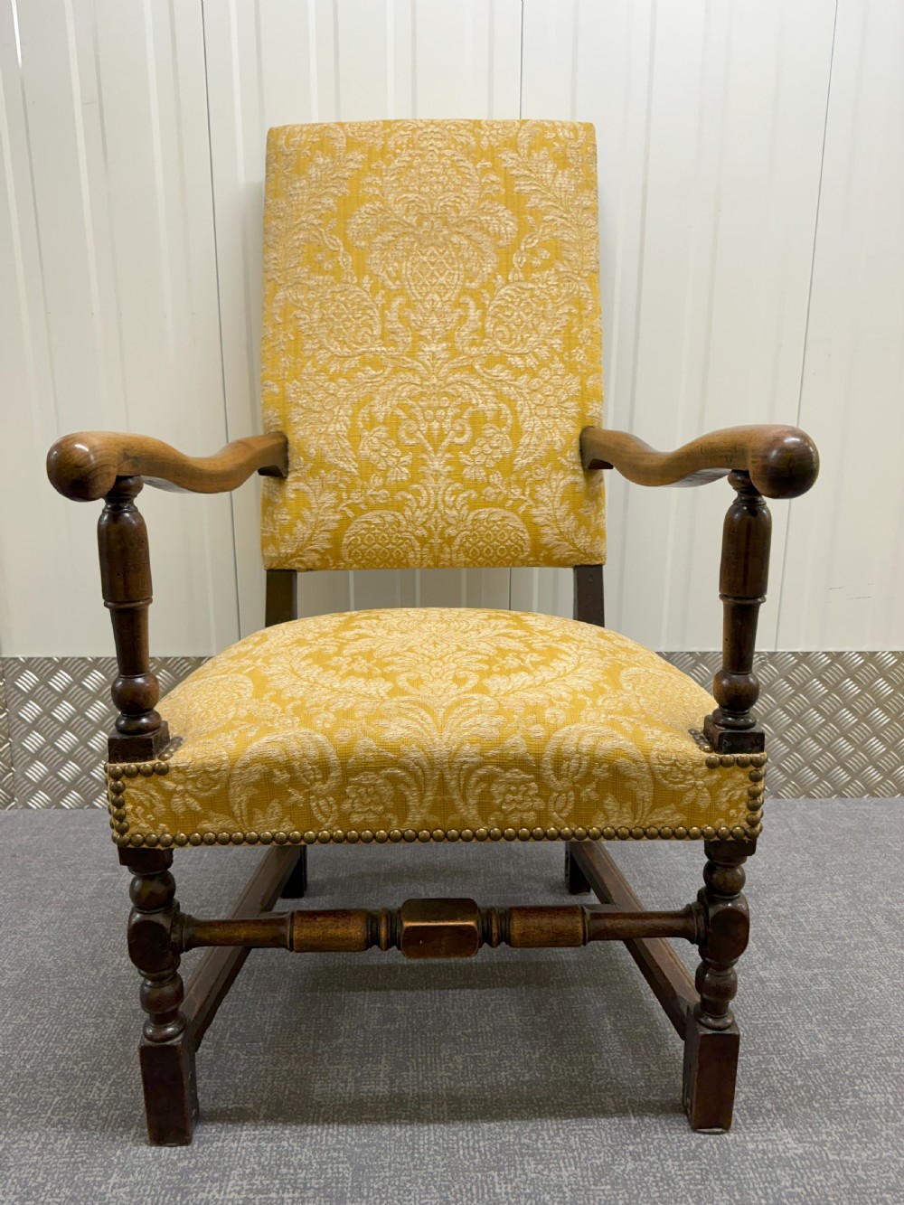 An antique Charles II style walnut elbow chair featuring turned wood legs, scrolled armrests, brass stud details, and yellow damask upholstery.