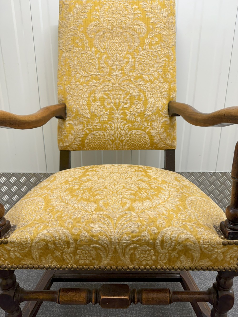 An antique Charles II style walnut elbow chair featuring turned wood legs, scrolled armrests, brass stud details, and yellow damask upholstery. View VI