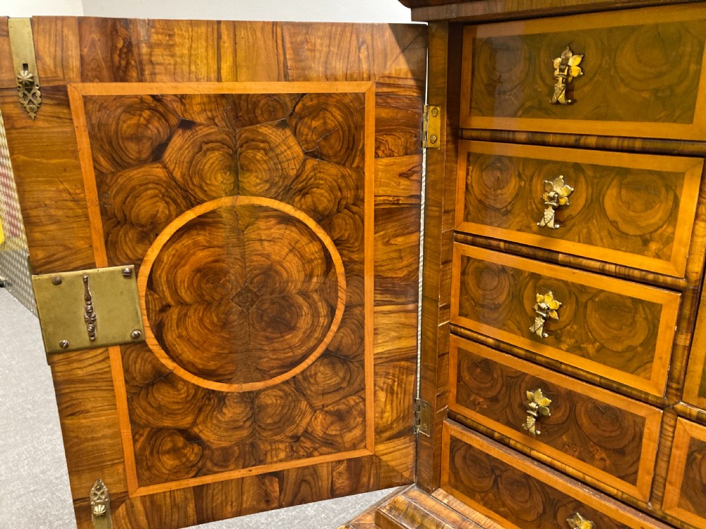 A William & Mary walnut cabinet on stand featuring door panels decorated with geometric oyster veneer stringing, resting on six barley-twist legs with a shaped stretcher base. View VI