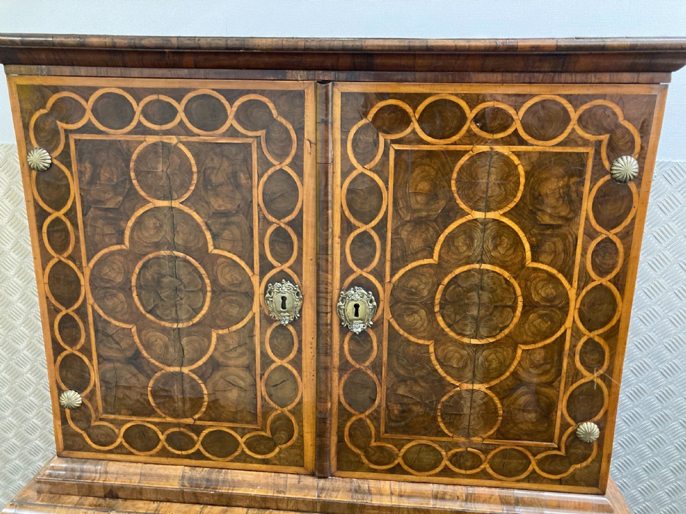 A William & Mary walnut cabinet on stand featuring door panels decorated with geometric oyster veneer stringing, resting on six barley-twist legs with a shaped stretcher base. View VII