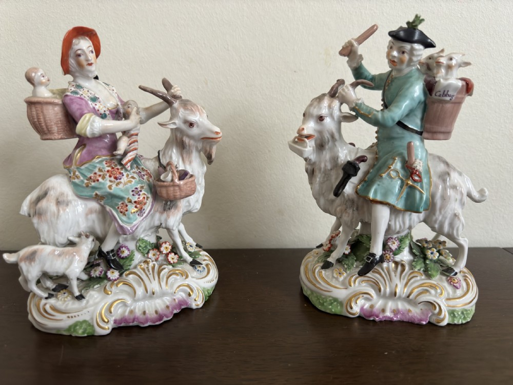 A pair of antique hand-painted porcelain figures depicting a man and woman riding goats decorated with floral details and rococo bases.