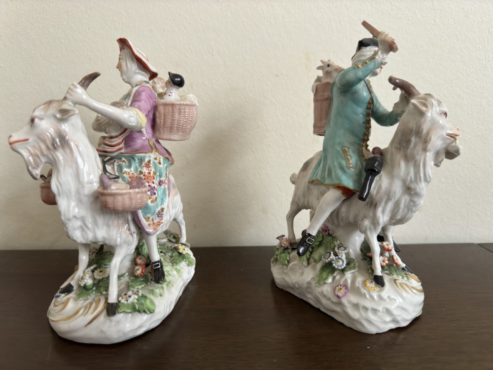 A pair of antique hand-painted porcelain figures depicting a man and woman riding goats decorated with floral details and rococo bases. View V