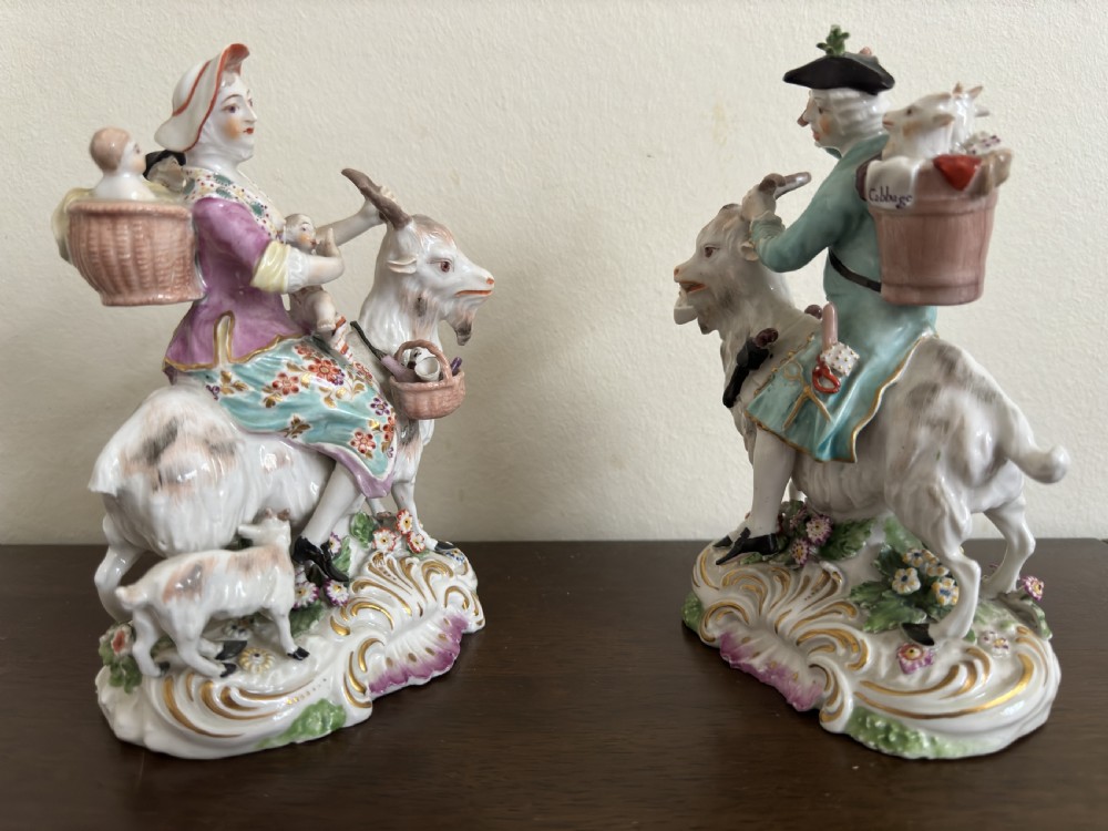 A pair of antique hand-painted porcelain figures depicting a man and woman riding goats decorated with floral details and rococo bases. View X