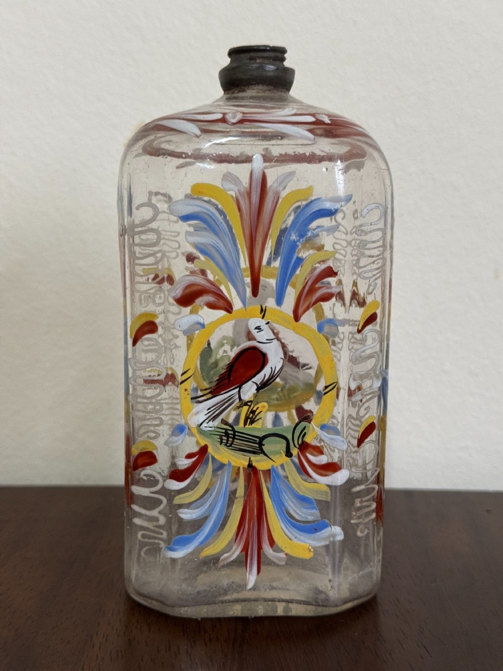 A rectangular clear glass bottle decorated with colourful hand-painted enamel floral motifs and a central bird emblem, fitted with a pewter screw top.