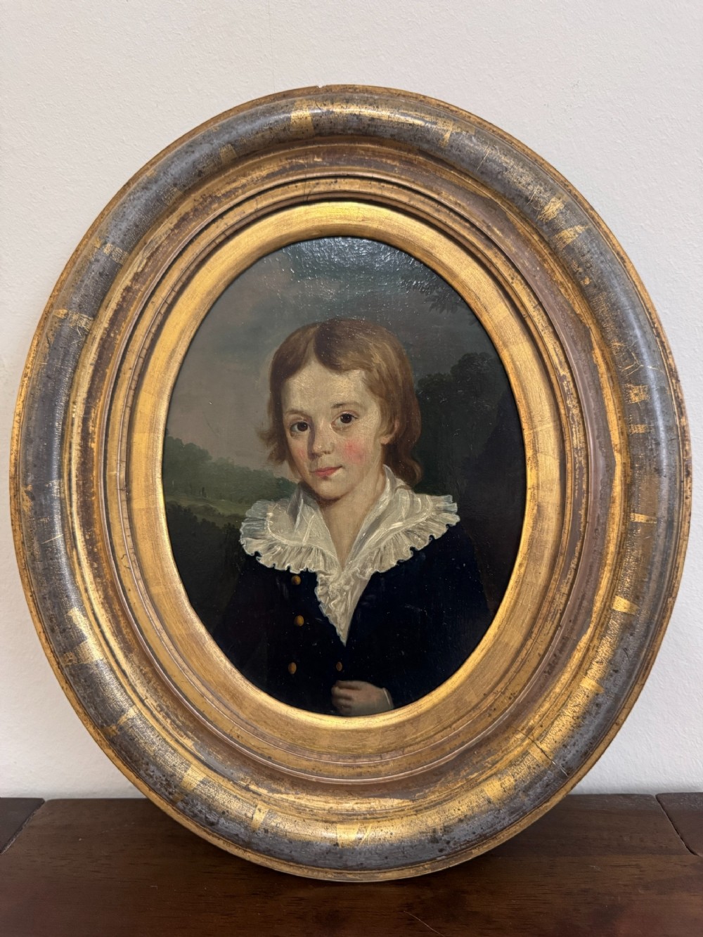 An 18th-century oval oil painting depicting a young boy with light brown hair wearing a white ruffled collar and dark blue jacket, set inside a carved giltwood frame.