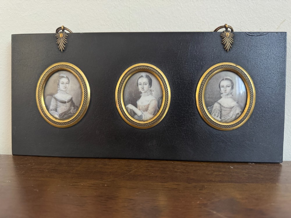A black rectangular panel displaying a set of three oval brass-framed 18th-century plumbago miniature portraits of women with decorative brass hanging mounts.