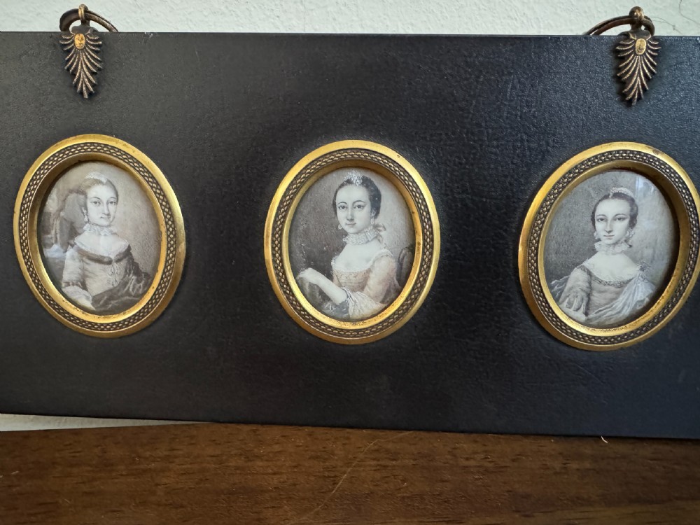 A black rectangular panel displaying a set of three oval brass-framed 18th-century plumbago miniature portraits of women with decorative brass hanging mounts. View II