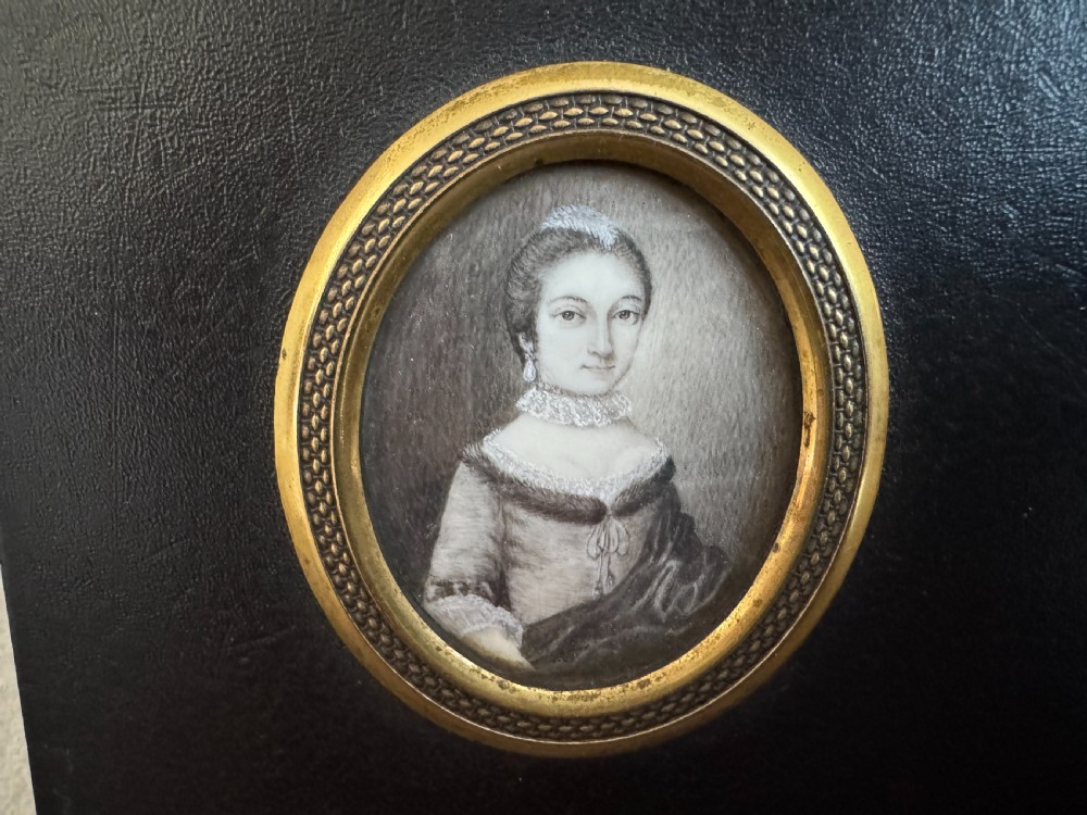 A black rectangular panel displaying a set of three oval brass-framed 18th-century plumbago miniature portraits of women with decorative brass hanging mounts. View III