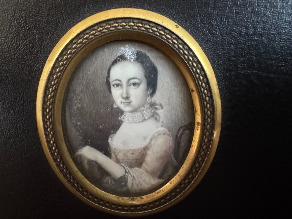 A black rectangular panel displaying a set of three oval brass-framed 18th-century plumbago miniature portraits of women with decorative brass hanging mounts. View IV