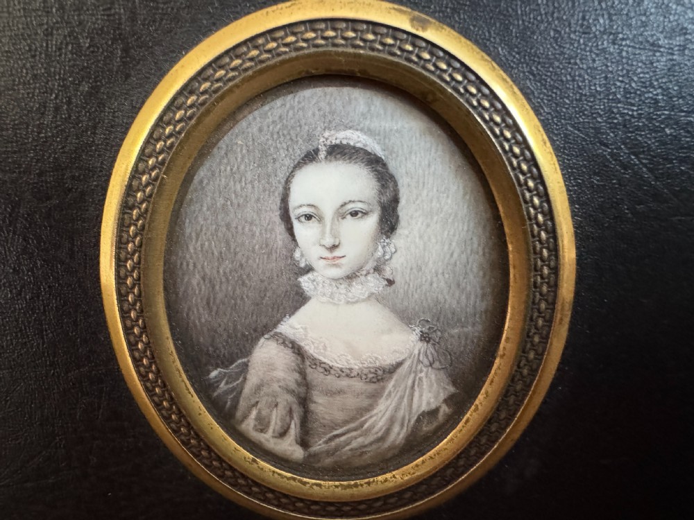A black rectangular panel displaying a set of three oval brass-framed 18th-century plumbago miniature portraits of women with decorative brass hanging mounts. View V