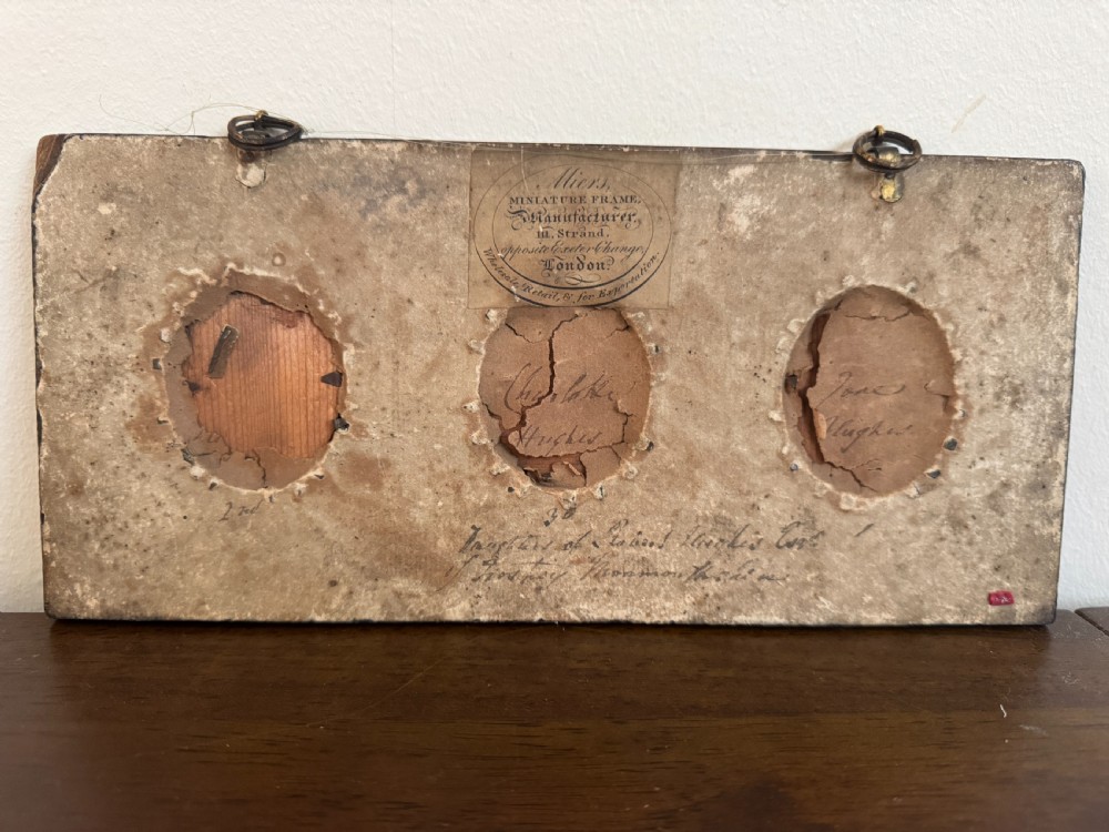 A black rectangular panel displaying a set of three oval brass-framed 18th-century plumbago miniature portraits of women with decorative brass hanging mounts. View VI