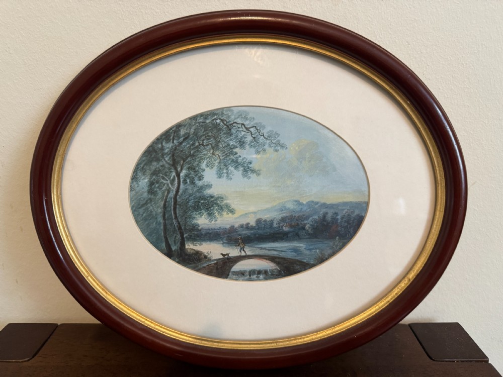 An oval 19th-century watercolour painting depicting a figure and dog crossing a stone bridge over a river, presented in a dark wood frame with a gold slip.