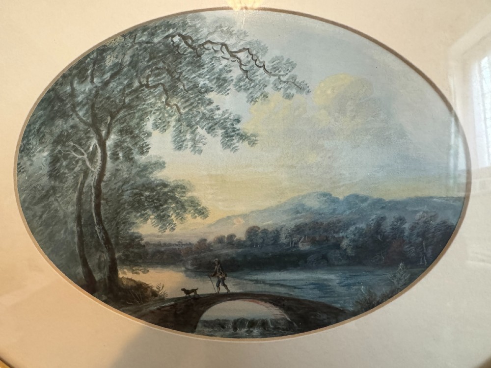 An oval 19th-century watercolour painting depicting a figure and dog crossing a stone bridge over a river, presented in a dark wood frame with a gold slip. View II