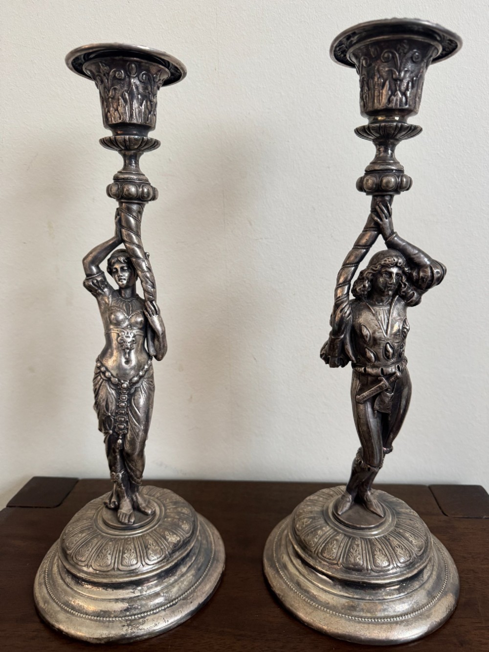 A pair of antique silver-plated candlesticks featuring detailed sculpted figures of a lady and a cavalier supporting sconces on rounded bases.